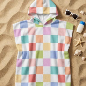 Puede incluir: Una toalla de playa con capucha con un patrón de tablero de ajedrez de colores pastel en tonos rosa, azul, verde, naranja y amarillo. La toalla es blanca con un diseño cuadrado repetido. Gafas de sol, protector solar y conchas marinas sobre la arena.