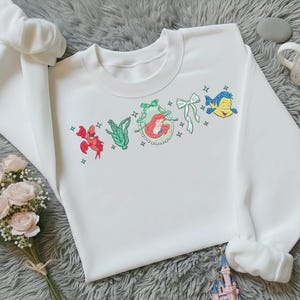 May include: White crewneck sweatshirt with embroidered cartoon characters from a popular animated film. The design includes a red crab, green seaweed, a framed image of a red-haired woman, a green bow, and a yellow and blue fish.