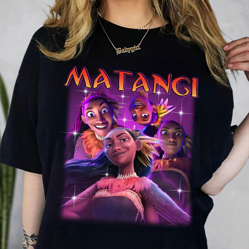 Op de afbeelding: Zwart T-shirt met een levendige afbeelding van geanimeerde personages en het woord "MATANGI" in oranje en geel. Het ontwerp bevat meerdere figuren met gedetailleerde gezichtsuitdrukkingen en kleurrijke kleding, tegen een paarse achtergrond.