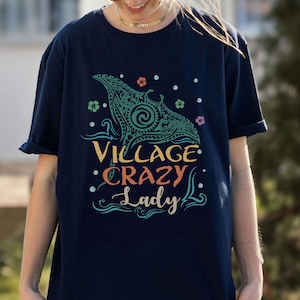 May include: Navy blue t-shirt with a teal and turquoise graphic design of a manta ray and the words "Village Crazy Lady" in colorful text. The design includes floral and dot accents. The shirt has short sleeves.