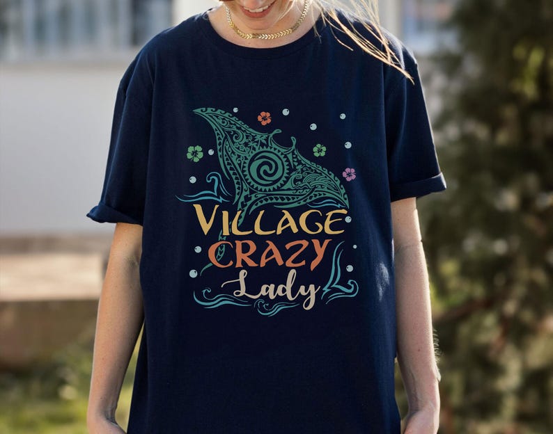 May include: Navy blue t-shirt with a teal and turquoise graphic design of a manta ray and the words "Village Crazy Lady" in colorful text. The design includes floral and dot accents. The shirt has short sleeves.