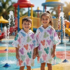 May include: Two children wearing white hooded beach towels with a colourful balloon design. The towels feature Mickey Mouse-shaped balloons in shades of pink, blue, green, and orange, with star accents. They are standing in front of a water fountain.