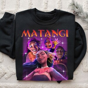 Op de afbeelding: Zwarte crewneck sweatshirt met een afbeelding van animatiefiguren en het woord "MATANGI" in oranje en geel. De afbeelding heeft een paarse achtergrond met sterren.