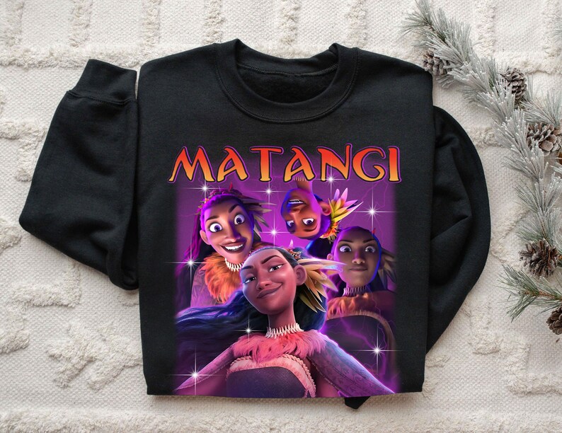 Op de afbeelding: Zwarte crewneck sweatshirt met een afbeelding van animatiefiguren en het woord "MATANGI" in oranje en geel. De afbeelding heeft een paarse achtergrond met sterren.