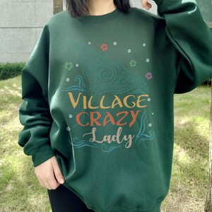 May include: A dark green sweatshirt with the text "VILLAGE CRAZY Lady" in a colorful, whimsical font. The design includes floral and paisley elements. The sweatshirt is a casual, comfortable style.