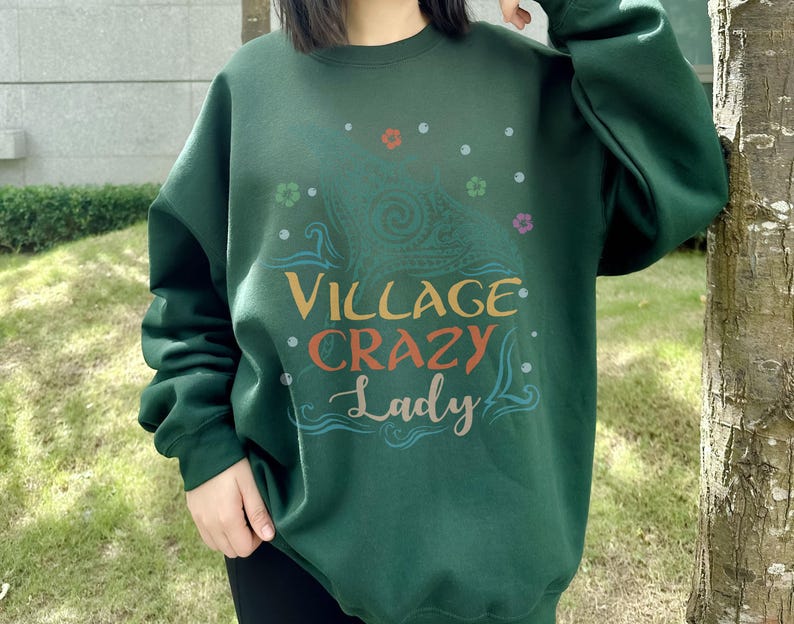 May include: A dark green sweatshirt with the text "VILLAGE CRAZY Lady" in a colorful, whimsical font. The design includes floral and paisley elements. The sweatshirt is a casual, comfortable style.