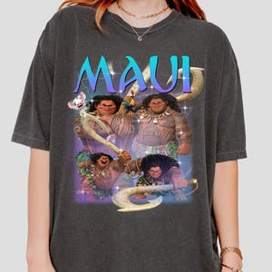 May include: Dark gray t-shirt featuring a graphic of the character Maui from the animated film Moana. The word "MAUI" is printed in a gradient blue and purple font above the character. The graphic includes images of Maui with his magical fish hook.