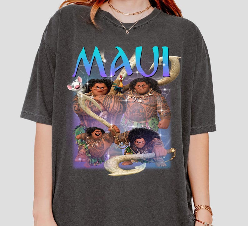 May include: Dark gray t-shirt featuring a graphic of the character Maui from the animated film Moana. The word "MAUI" is printed in a gradient blue and purple font above the character. The graphic includes images of Maui with his magical fish hook.