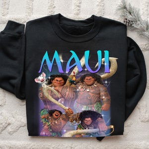 May include: Black sweatshirt featuring a graphic of the character Maui from the animated film Moana. The word "MAUI" is displayed in a gradient blue and purple font. The design includes illustrations of Maui with his tattoos and a pig.