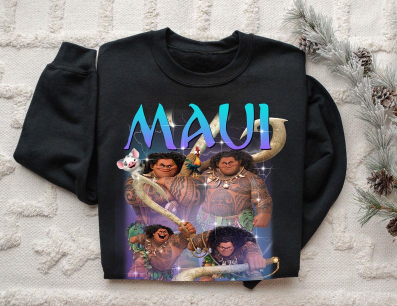 May include: Black sweatshirt featuring a graphic of the character Maui from the animated film Moana. The word "MAUI" is displayed in a gradient blue and purple font. The design includes illustrations of Maui with his tattoos and a pig.
