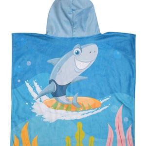 May include: A hooded, blue shark-themed beach towel. The towel features a cartoon shark riding a surfboard on a blue background with white waves and yellow and pink seaweed. The hood is a lighter shade of blue.