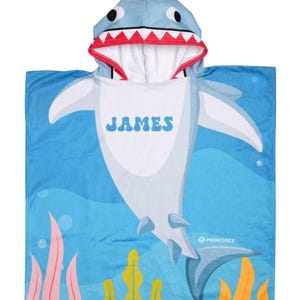 May include: A blue shark-themed hooded towel with the name "JAMES" printed on the front. The towel features a shark design with a white belly, gray fins, and a hood shaped like a shark's head with red and white teeth. The background is blue with aquatic plants.