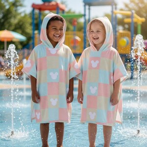 May include: Two children wearing matching hooded ponchos with a pastel checkered pattern and white Mickey Mouse silhouettes. They are standing in a splash pad with water fountains, smiling and laughing. The ponchos are light blue, pink, and yellow.