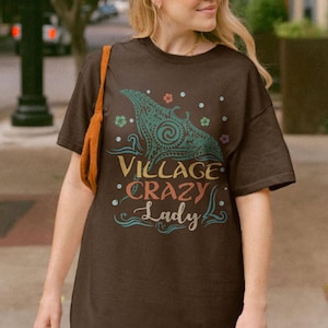 May include: A brown t-shirt with the text "Village Crazy Lady" in a colorful, whimsical font. The shirt features a teal and yellow graphic design of a manta ray. The shirt is short-sleeved.