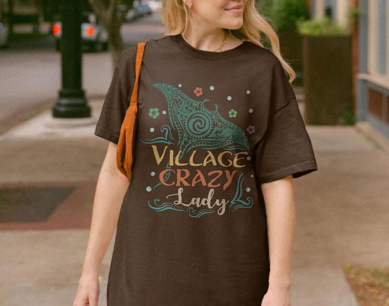 May include: A brown t-shirt with the text "Village Crazy Lady" in a colorful, whimsical font. The shirt features a teal and yellow graphic design of a manta ray. The shirt is short-sleeved.
