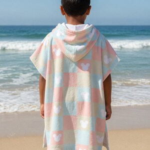May include: A child's hooded beach towel in pastel pink, blue, and cream, featuring a checkered design with white Mickey Mouse silhouettes. The child is on a sandy beach, with the sea visible in the background.