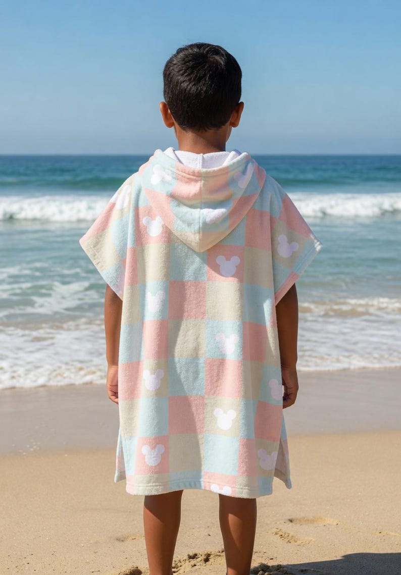 May include: A child's hooded beach towel in pastel pink, blue, and cream, featuring a checkered design with white Mickey Mouse silhouettes. The child is on a sandy beach, with the sea visible in the background.