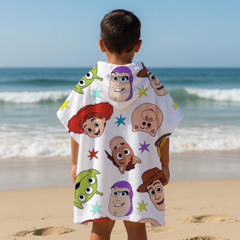May include: A white hooded beach towel featuring colorful cartoon character faces. The towel is patterned with images of Buzz Lightyear, Woody, and other characters. The child is standing on a sandy beach with the ocean in the background.