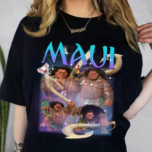 May include: Black t-shirt featuring a graphic of the character Maui from the animated film Moana. The word "MAUI" is displayed in large, blue and purple letters. The image includes illustrations of Maui with his magical fish hook and other characters.