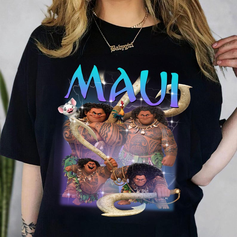 May include: Black t-shirt featuring a graphic of the character Maui from the animated film Moana. The word "MAUI" is displayed in large, blue and purple letters. The image includes illustrations of Maui with his magical fish hook and other characters.