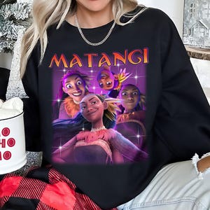 Op de afbeelding: Zwarte sweatshirt met een afbeelding van geanimeerde personages en het woord "MATANGI" in oranje. Het ontwerp toont vier personages met verschillende uitdrukkingen. Een witte mok met "HO HO HO" in rood is zichtbaar.