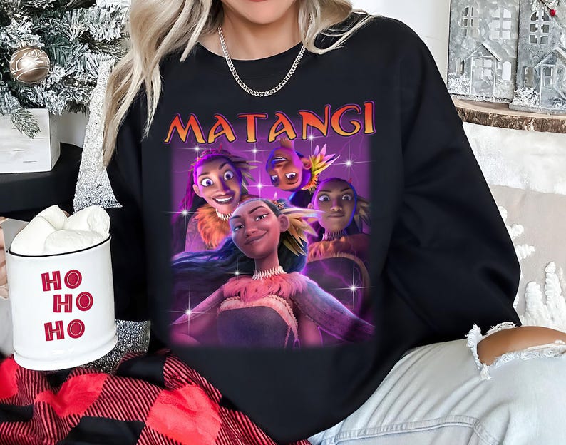 Op de afbeelding: Zwarte sweatshirt met een afbeelding van geanimeerde personages en het woord "MATANGI" in oranje. Het ontwerp toont vier personages met verschillende uitdrukkingen. Een witte mok met "HO HO HO" in rood is zichtbaar.