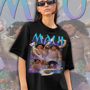 May include: Black t-shirt featuring the word "MAUI" in teal and purple, with a graphic of the character Maui from the animated film Moana. The design includes images of Maui with his magical fish hook and other characters from the movie.