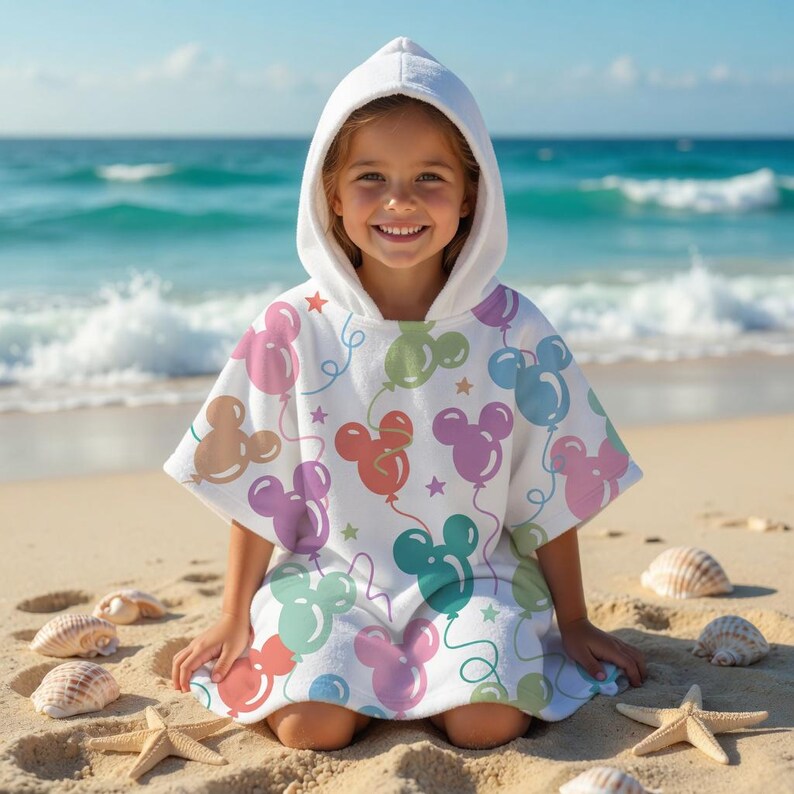 May include: A white hooded beach towel with a colourful Mickey Mouse balloon pattern. The towel has a hood and is designed for children. The background shows a sandy beach, seashells, and the ocean.