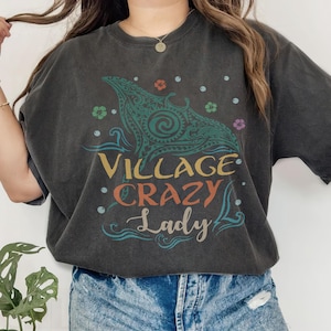 May include: Dark gray t-shirt with a colorful graphic. The design features a stylized sea creature with the words "VILLAGE CRAZY Lady" in a mix of fonts and colors. Floral and dot accents surround the central image.