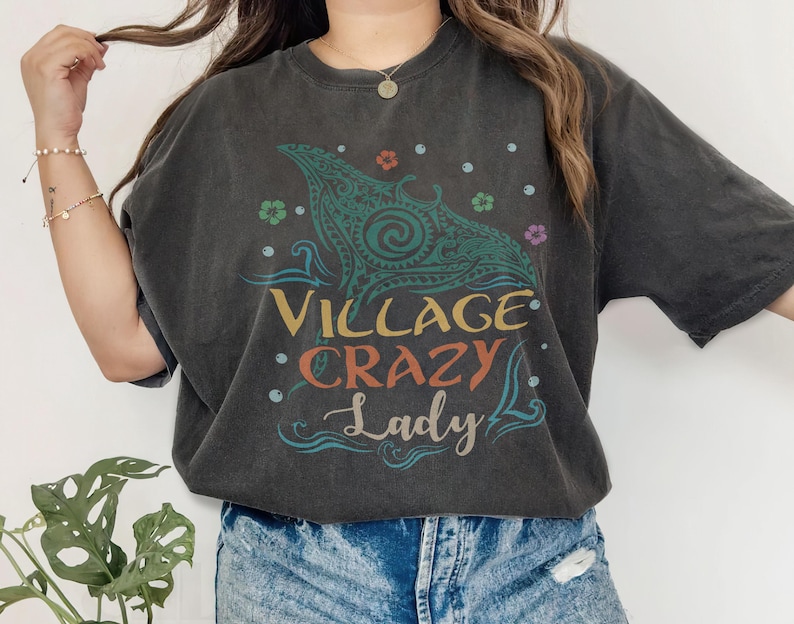 May include: Dark gray t-shirt with a colorful graphic. The design features a stylized sea creature with the words "VILLAGE CRAZY Lady" in a mix of fonts and colors. Floral and dot accents surround the central image.