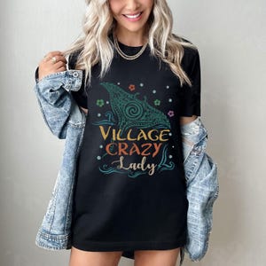 May include: Black t-shirt with the text "Village Crazy Lady" in orange, yellow, and teal. The shirt features a teal and green graphic design. The shirt is worn with a denim jacket.
