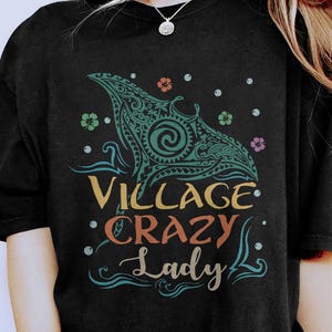 May include: Black t-shirt featuring a teal and turquoise manta ray design with tribal patterns. The text "Village Crazy Lady" is in yellow, orange, and teal. Small floral and dot accents surround the design.