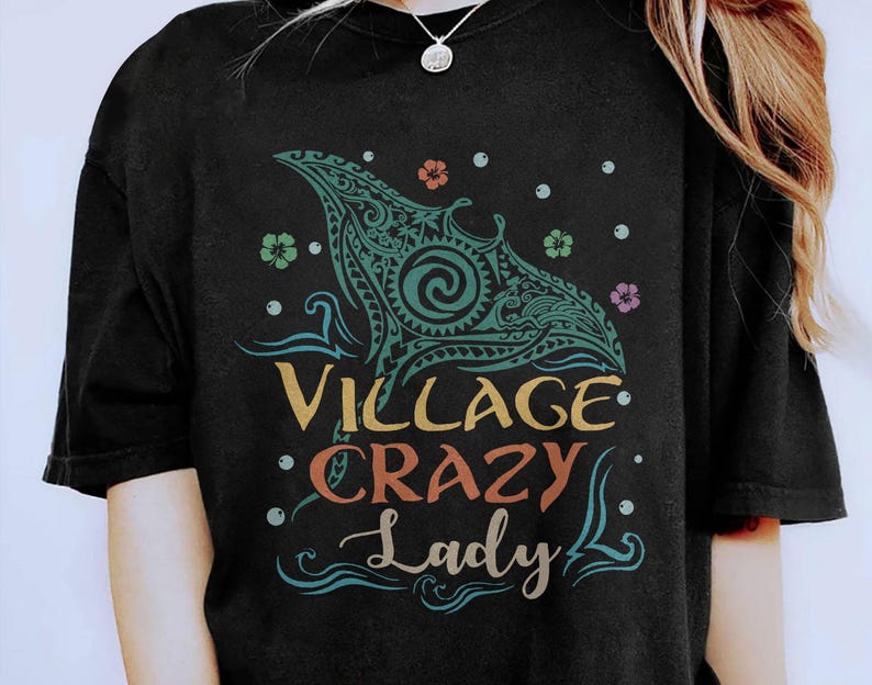 May include: Black t-shirt featuring a teal and turquoise manta ray design with tribal patterns. The text "Village Crazy Lady" is in yellow, orange, and teal. Small floral and dot accents surround the design.