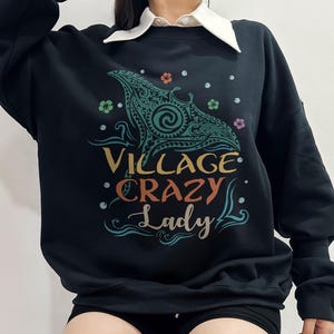 May include: Black sweatshirt with a teal and green graphic of a manta ray and the words "VILLAGE CRAZY Lady" in colorful text. The sweatshirt has a crew neck and long sleeves. The design includes floral and wave elements.