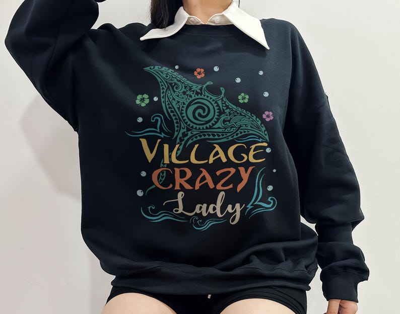 May include: Black sweatshirt with a teal and green graphic of a manta ray and the words "VILLAGE CRAZY Lady" in colorful text. The sweatshirt has a crew neck and long sleeves. The design includes floral and wave elements.