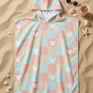 May include: A pastel-coloured hooded beach towel with a chequered pattern of pink, blue, and beige squares. White Mickey Mouse head silhouettes are spaced across the towel. The towel is on sand, with sunglasses and a drink nearby.
