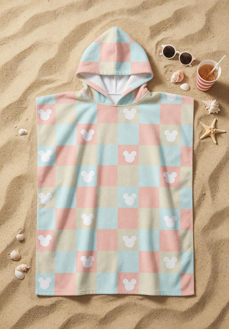 May include: A pastel-coloured hooded beach towel with a chequered pattern of pink, blue, and beige squares. White Mickey Mouse head silhouettes are spaced across the towel. The towel is on sand, with sunglasses and a drink nearby.