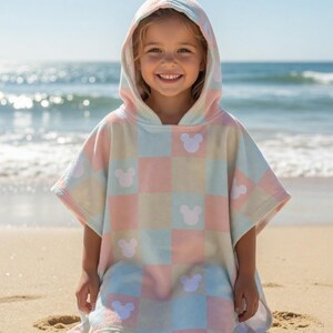 May include: A pastel-coloured hooded beach towel with a checkered pattern and white Mickey Mouse silhouettes. A child wears the towel on a sandy beach, with the sea visible in the background.