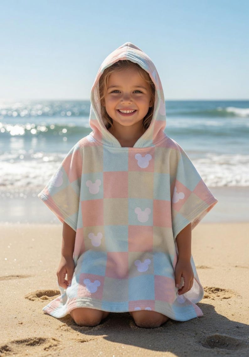 May include: A pastel-coloured hooded beach towel with a checkered pattern and white Mickey Mouse silhouettes. A child wears the towel on a sandy beach, with the sea visible in the background.