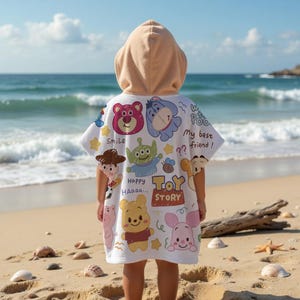 May include: A white hooded beach towel featuring various Toy Story characters, including Woody, Buzz Lightyear, and Winnie the Pooh. The towel has a tan hood and is being worn on a sandy beach with the ocean in the background.
