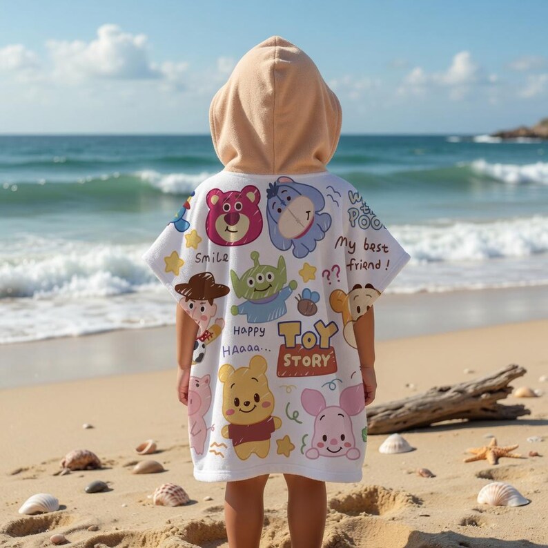 May include: A white hooded beach towel featuring various Toy Story characters, including Woody, Buzz Lightyear, and Winnie the Pooh. The towel has a tan hood and is being worn on a sandy beach with the ocean in the background.