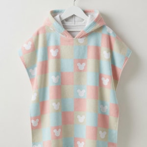 May include: A pastel-coloured hooded poncho towel with a checkered design in pink, blue, and beige. White Mickey Mouse head silhouettes are spaced across the squares. The towel is hanging on a white hanger.