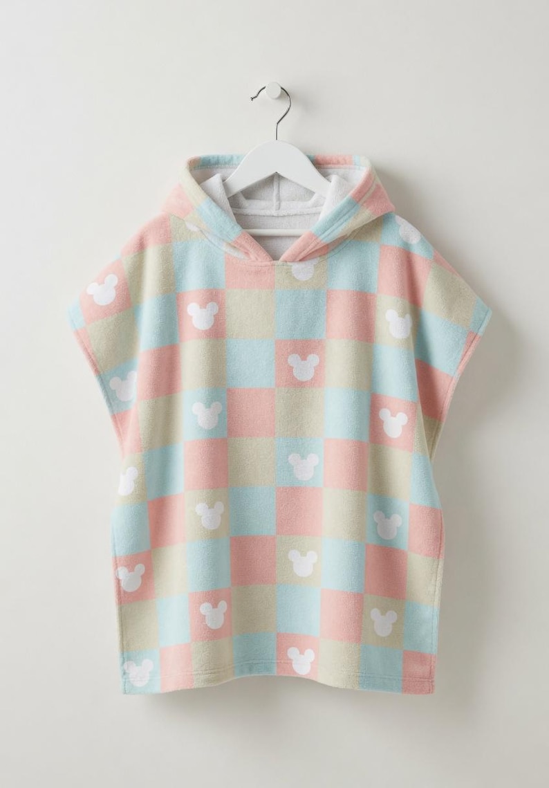 May include: A pastel-coloured hooded poncho towel with a checkered design in pink, blue, and beige. White Mickey Mouse head silhouettes are spaced across the squares. The towel is hanging on a white hanger.
