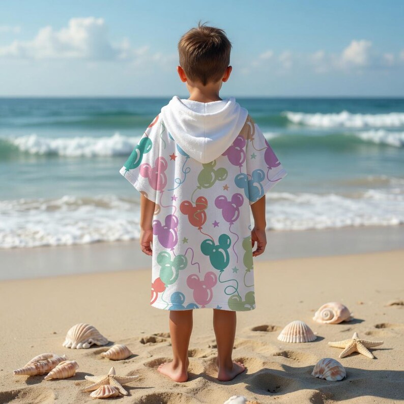 May include: A white hooded beach towel with a colourful balloon design featuring Mickey Mouse ears. The towel is worn by a child on a sandy beach with seashells and the ocean in the background.