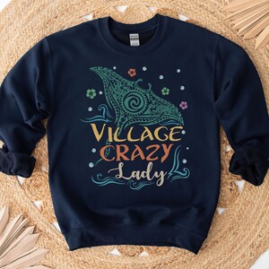 May include: Navy blue sweatshirt with the text "VILLAGE CRAZY Lady" in colorful lettering. The design includes a teal manta ray graphic with floral and dot accents. The sweatshirt is displayed on a woven, tan surface.
