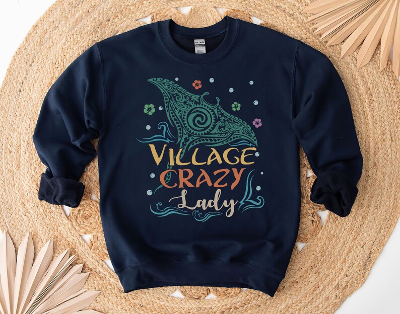 May include: Navy blue sweatshirt with the text "VILLAGE CRAZY Lady" in colorful lettering. The design includes a teal manta ray graphic with floral and dot accents. The sweatshirt is displayed on a woven, tan surface.