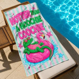 Könnte beinhalten: Strandtuch mit rosa und grün gestreiftem Hintergrund. Der Text "HOTTER THAN A HOOCHIE COOCHIE" ist in Rosa und Grün gehalten. Ein Cartoon-Alligator mit Sonnenbrille und ein rosa Flamingo befinden sich auf einem rosa Schlauchboot im Wasser.