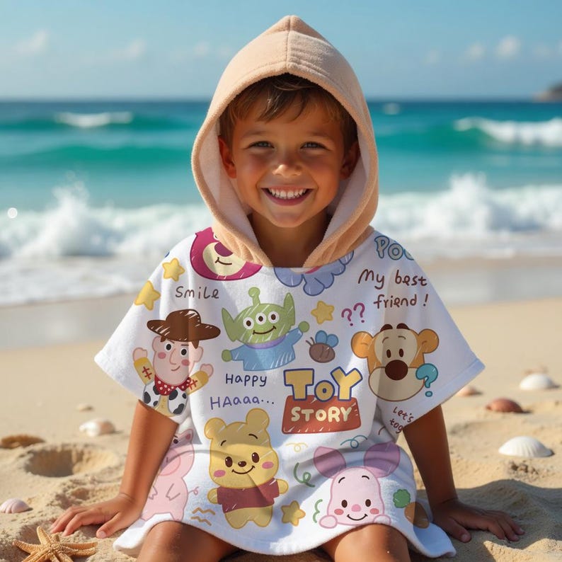 May include: A child wearing a white hooded beach towel with colorful Toy Story character graphics. The towel features illustrations of Woody, an alien, Winnie the Pooh, and Piglet, with the words "Toy Story" and other phrases.