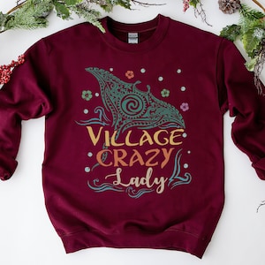 May include: Burgundy sweatshirt with the text "Village Crazy Lady" in a colorful design. The graphic includes a teal and gold tribal-style whale illustration, flowers, and dots. A cozy, casual top.