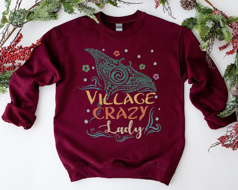May include: Burgundy sweatshirt with the text "Village Crazy Lady" in a colorful design. The graphic includes a teal and gold tribal-style whale illustration, flowers, and dots. A cozy, casual top.